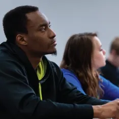 A diverse group of students sitting at desks in a classroom, listening to the teacher and taking notes.